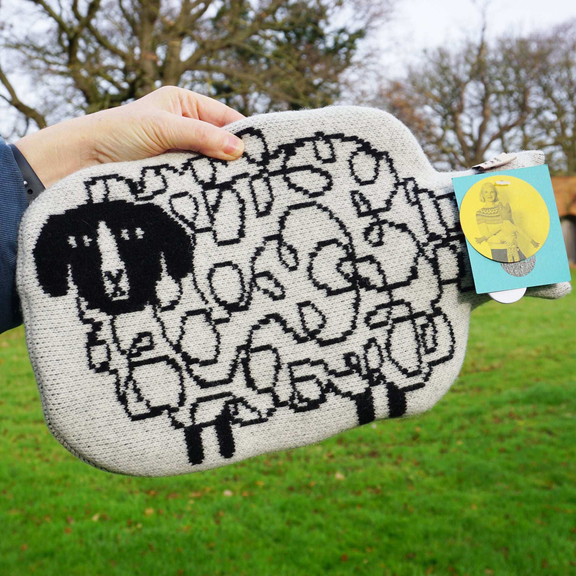 A hand holding a grey wool hot water bottle that features a black knitted sheep pattern and swing tags.