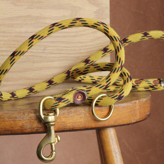 A yellow dog rope lead with a solid brass clip and copper rivets, made from recycled climbing rope and oak-tanned leather, displayed on a wooden stool.