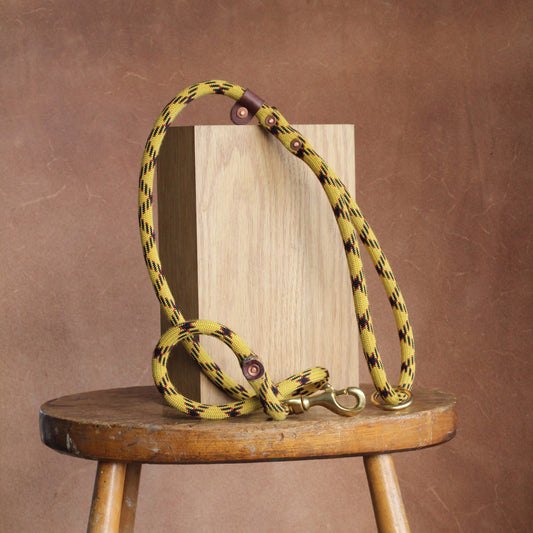 A yellow dog rope lead with a solid brass clip and copper rivets, made from recycled climbing rope and oak-tanned leather, displayed on a wooden stool.