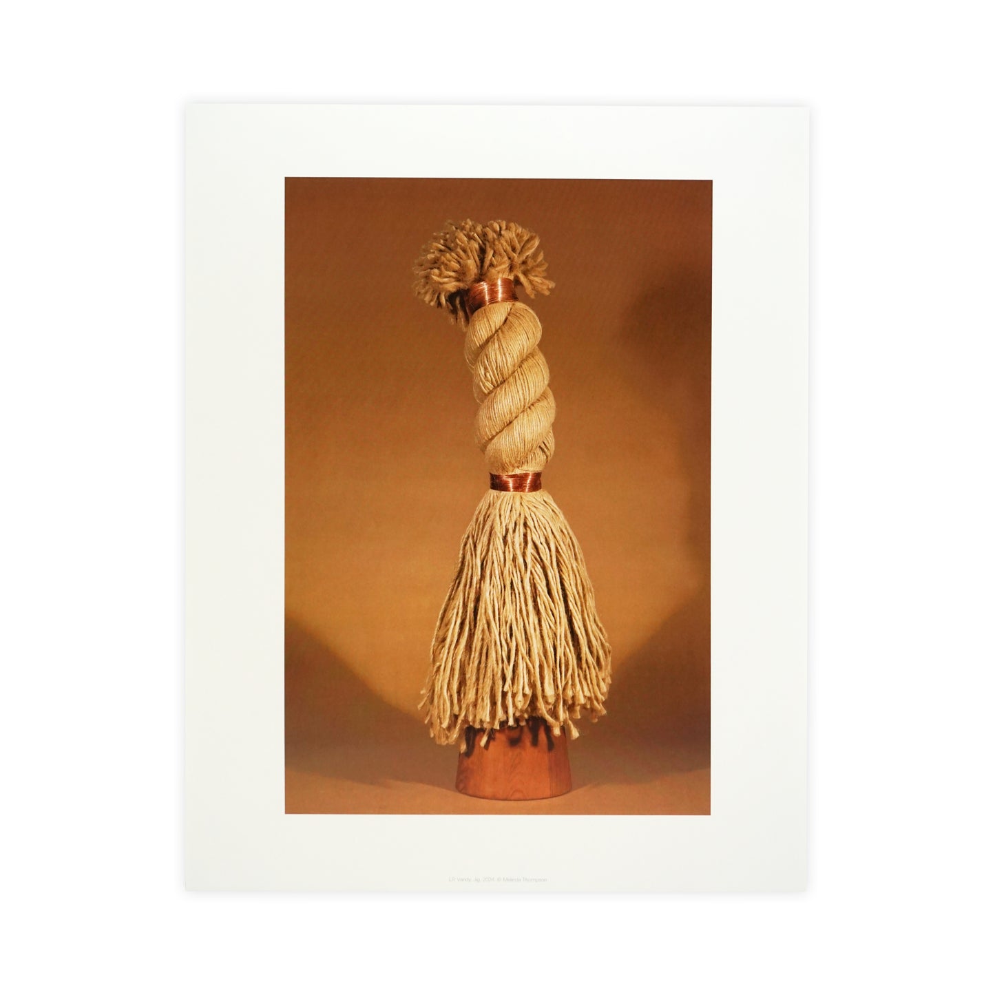 A decorative object resembling a tassel, featuring a twisted top and long, flowing strands. The item is displayed against a warm, neutral background, highlighting its texture and craftsmanship. The overall tone is earthy and inviting.