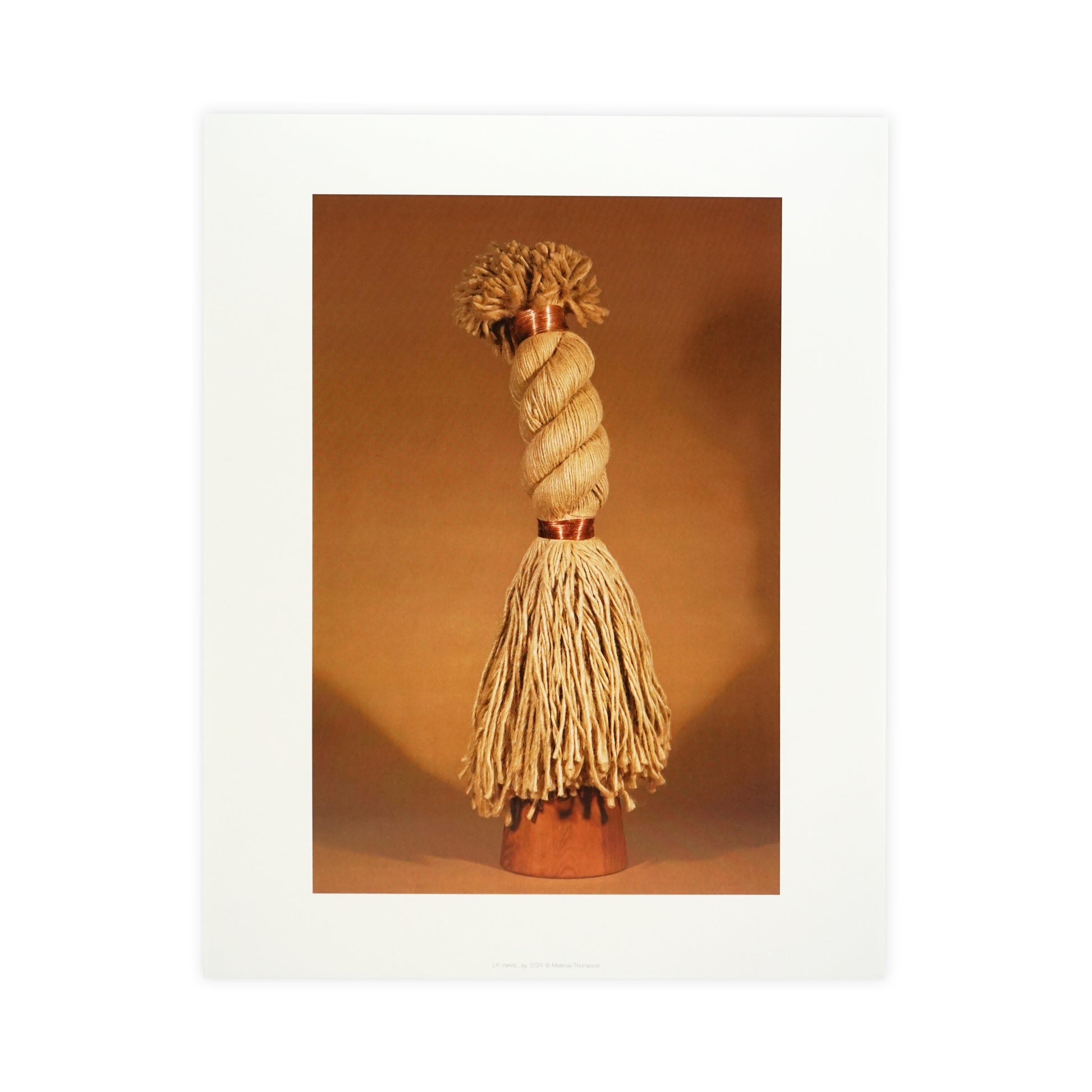 A decorative object resembling a tassel, featuring a twisted top and long, flowing strands. The item is displayed against a warm, neutral background, highlighting its texture and craftsmanship. The overall tone is earthy and inviting.