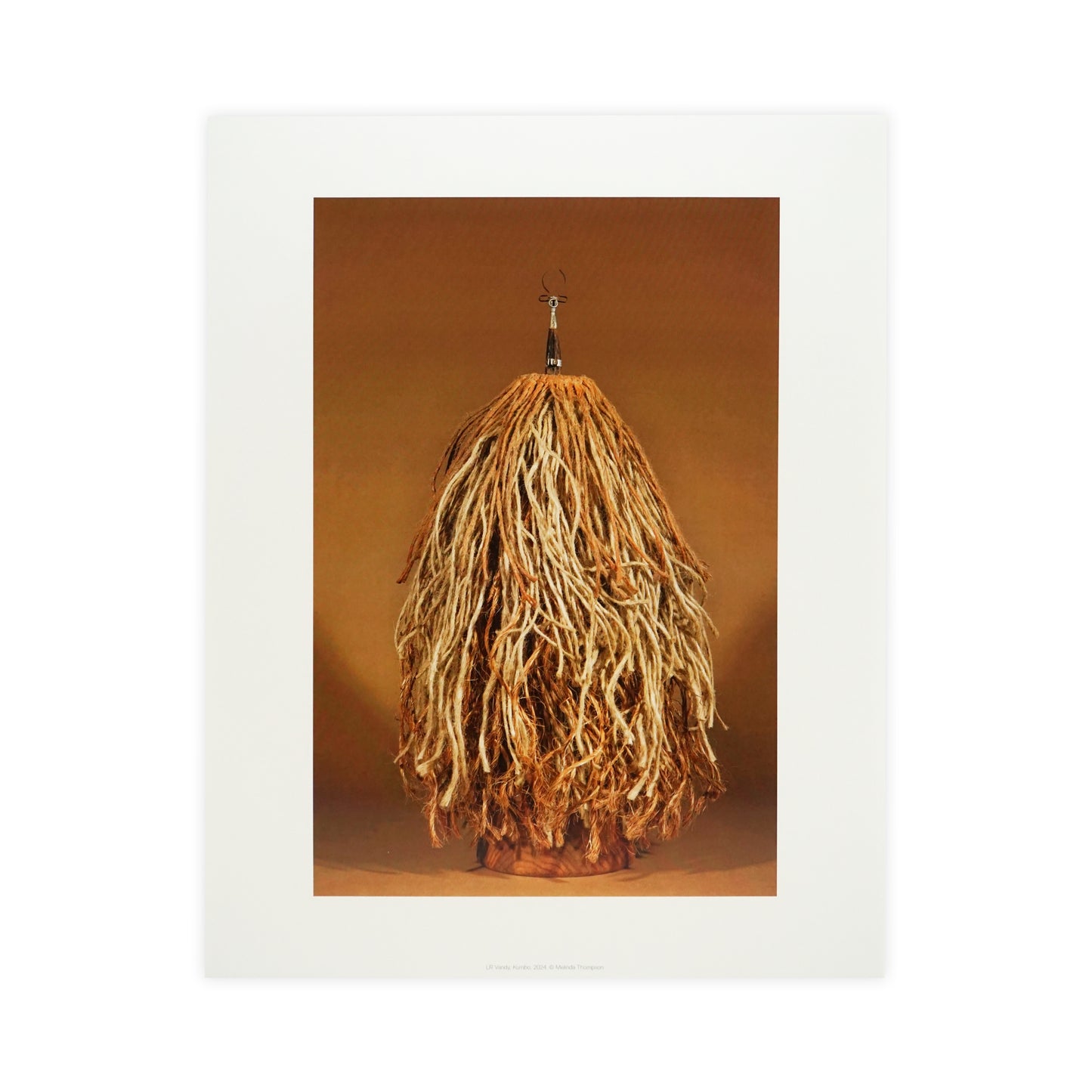 A large, textured sculpture resembling a mop or a bundle of long, thin strands hangs against a warm, neutral background. The piece features a mix of earthy tones, creating a natural and organic appearance.