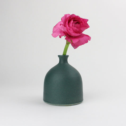 A hand thrown earthenware grey/green vase with a speckled matt glaze featuring a single stem of a pink rose inside.