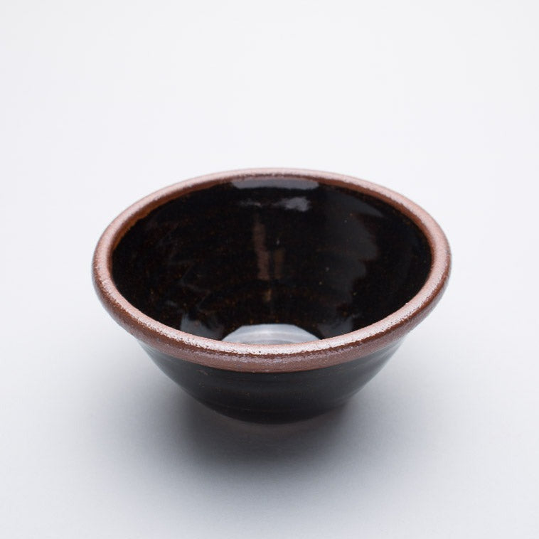 A small stoneware ceramic bowl with a black and brown hue and a dark brown rim.