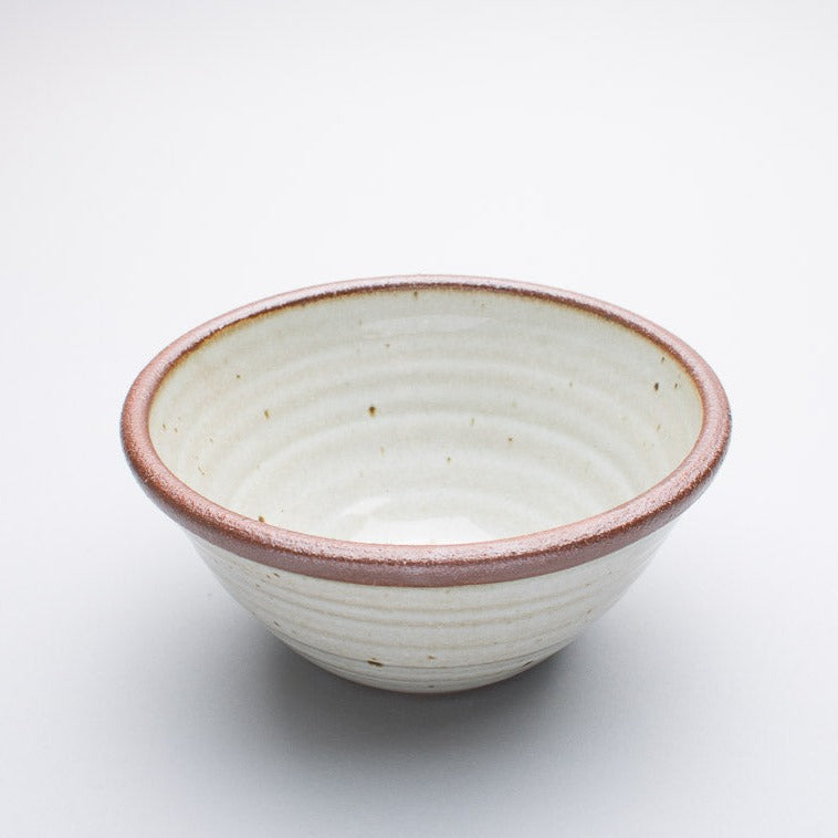 A medium stoneware ceramic bowl with a cream hue and a dark brown rim.