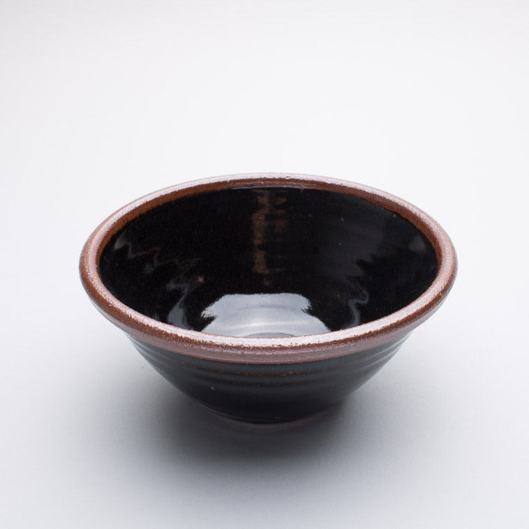 A medium stoneware ceramic bowl with a black and brown hue and a dark brown rim.