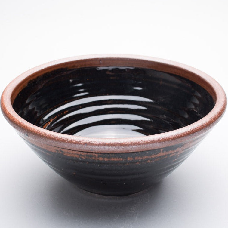 A large stoneware ceramic bowl with a black and brown hue and a dark brown rim.