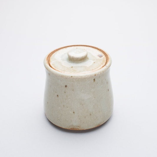 A beige ceramic honey jar with a lid, made of stoneware clay, with a creamy white glaze that may have a hint of blue.