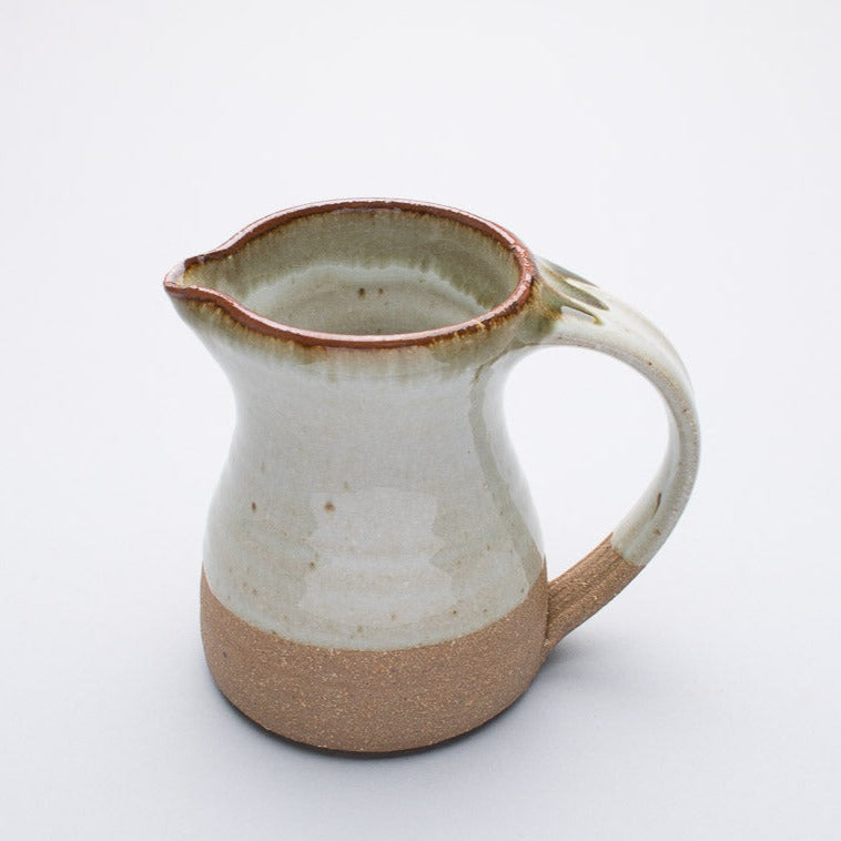 A small ceramic jug with a two-tone colour, primarily a creamish hue with a darker brown at the rim and handle.
