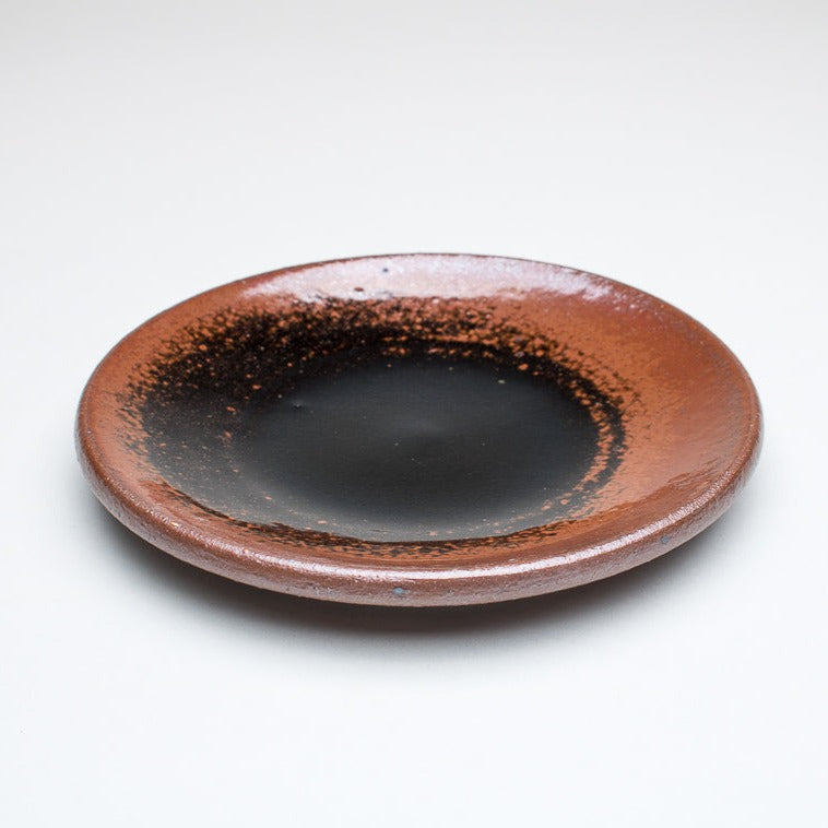 A ceramic plate with a browny black centre and a greenish-brown rim, with variations in the glaze that give it a unique texture.