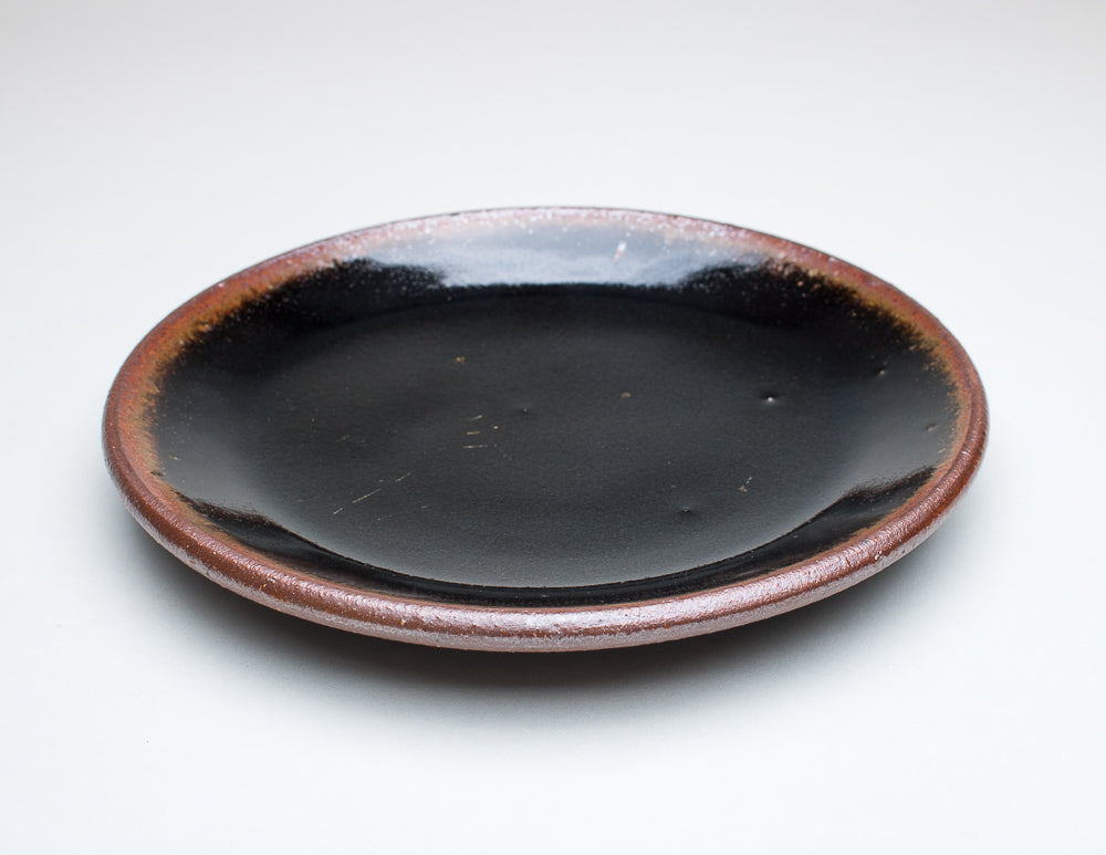 A ceramic plate with a black centre and a greenish-brown rim, with variations in the glaze that give it a unique texture.