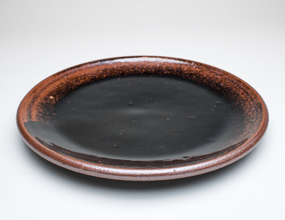 A ceramic plate with a black centre and a greenish-brown rim, with variations in the glaze that give it a unique texture.