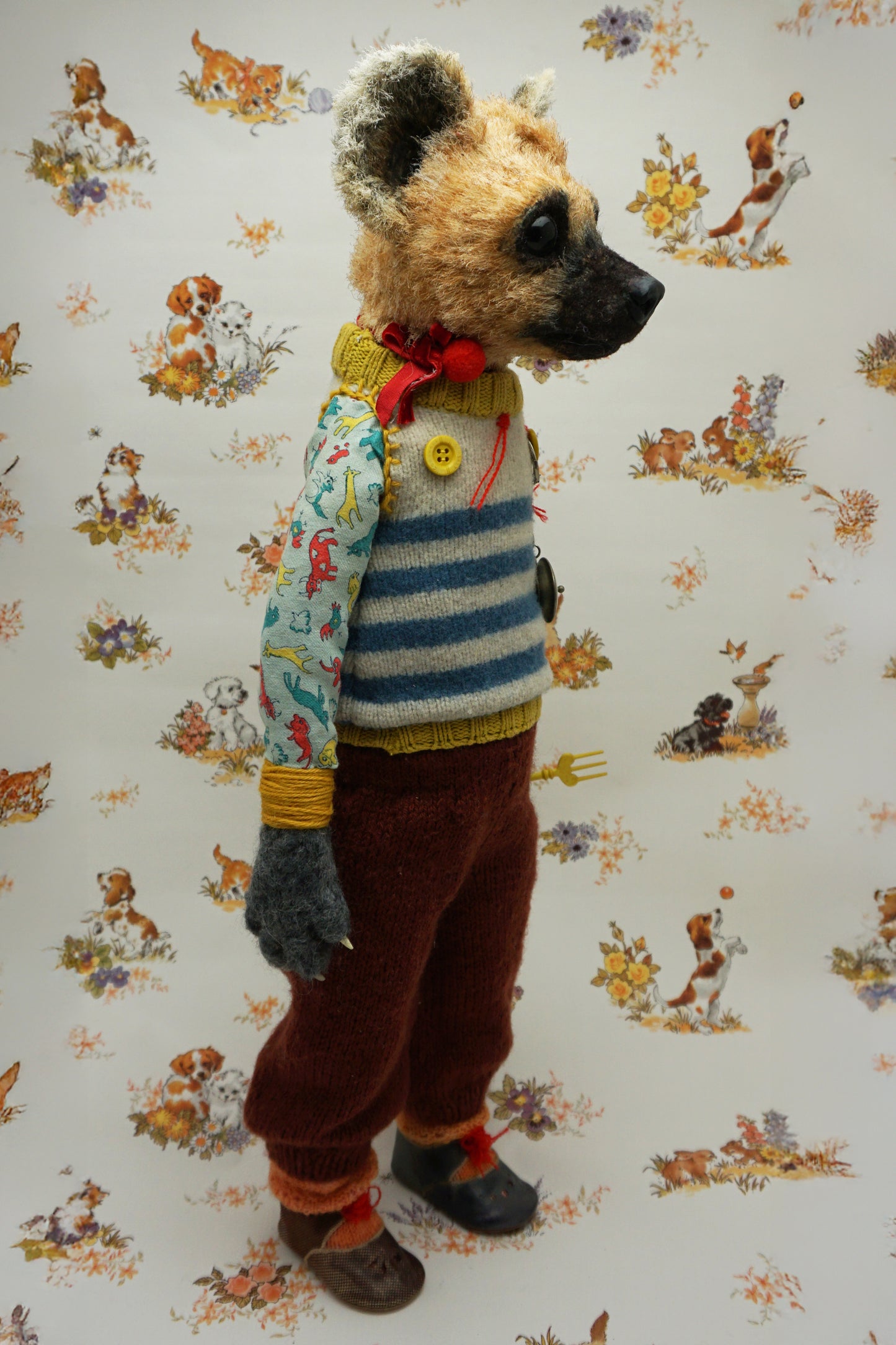 A handmade plush doll resembling a hyena stands against a floral background. It wears a striped jumper, patterned shirt, and red trousers. The doll has a fork accessory and button details, exuding a whimsical, vintage charm.