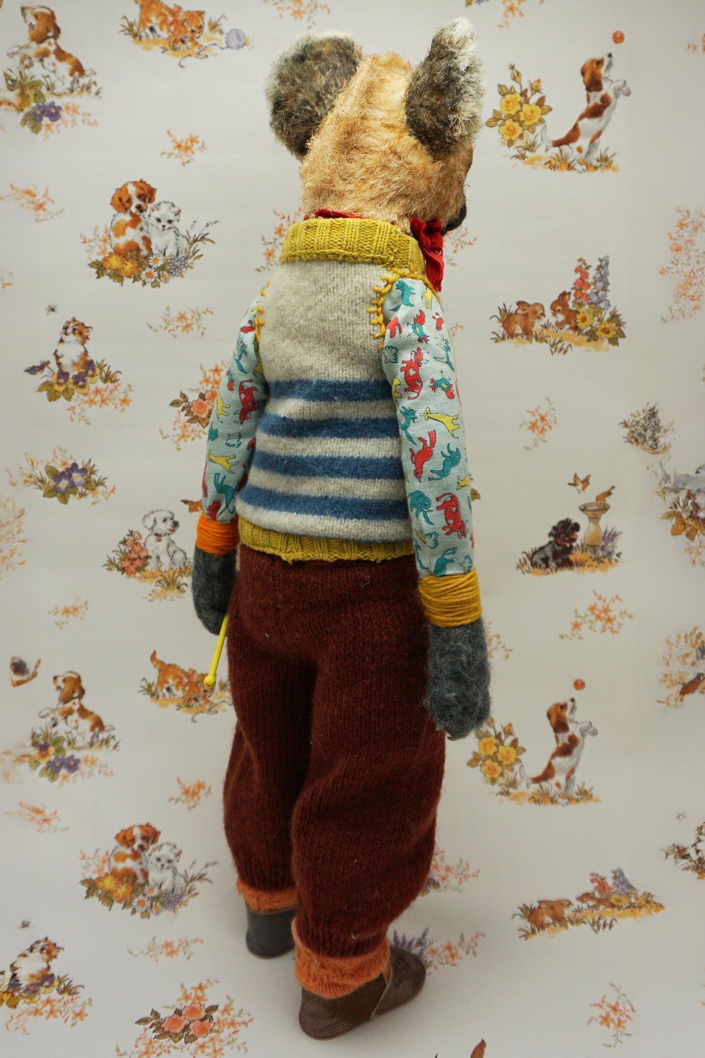 A handmade plush doll resembling a hyena stands against a floral background. It wears a striped jumper, patterned shirt, and red trousers. The doll has a fork accessory and button details, exuding a whimsical, vintage charm.