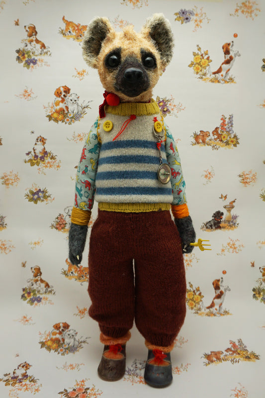 A handmade plush doll resembling a hyena stands against a floral background. It wears a striped jumper, patterned shirt, and red trousers. The doll has a fork accessory and button details, exuding a whimsical, vintage charm.