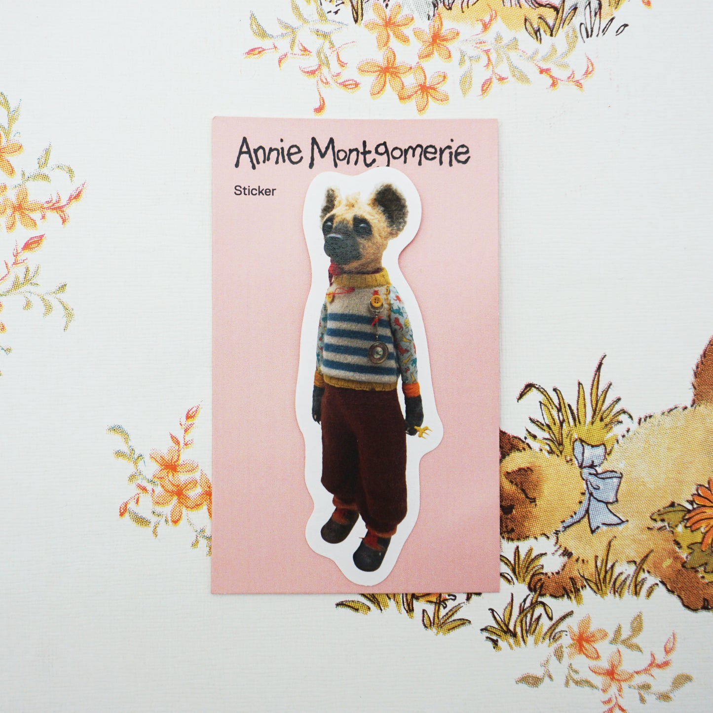 A die-cut sticker featuring a hyena wearing a striped sweater and brown pants, placed on a pink backing card labelled 'Annie Montgomerie Sticker'. The card is photographed on a white background with a decorative floral print.