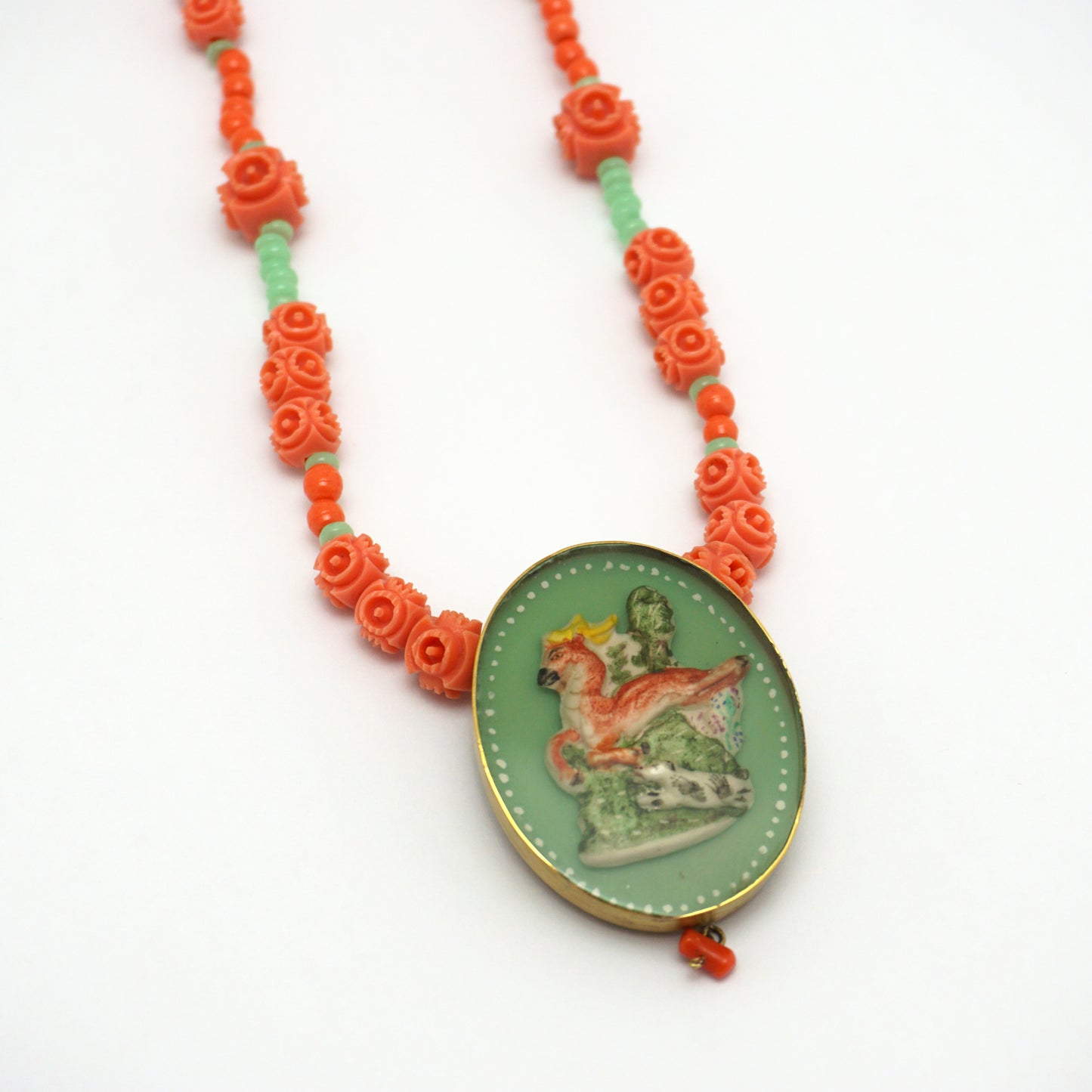 Necklace with an oval hand painted pendant insert that features an animal design on a green background. The necklace chain is made up of orange and green rose shapes Bakelite beads. 