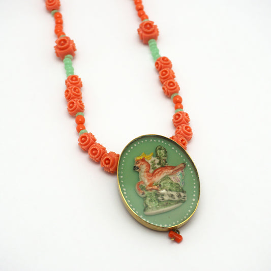 Necklace with an oval hand painted pendant insert that features an animal design on a green background. The necklace chain is made up of orange and green rose shapes Bakelite beads. 