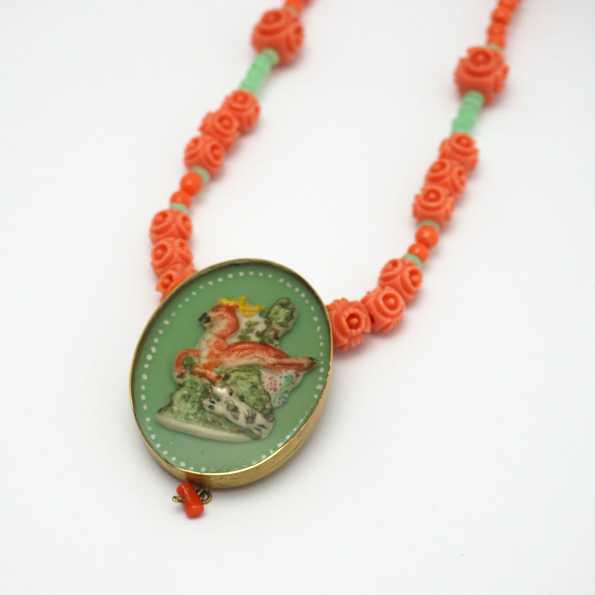 Necklace with an oval hand painted pendant insert that features an animal design on a green background. The necklace chain is made up of orange and green rose shapes Bakelite beads. 