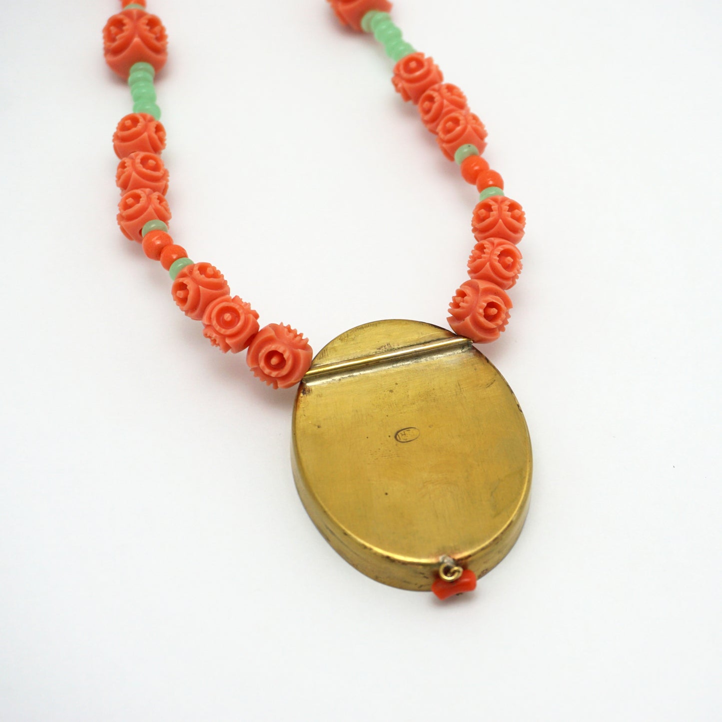 The reverse of a necklace with an oval pendant. The back of the charm is smooth and shiny. The necklace chain is made up of orange and green rose shapes Bakelite beads. 