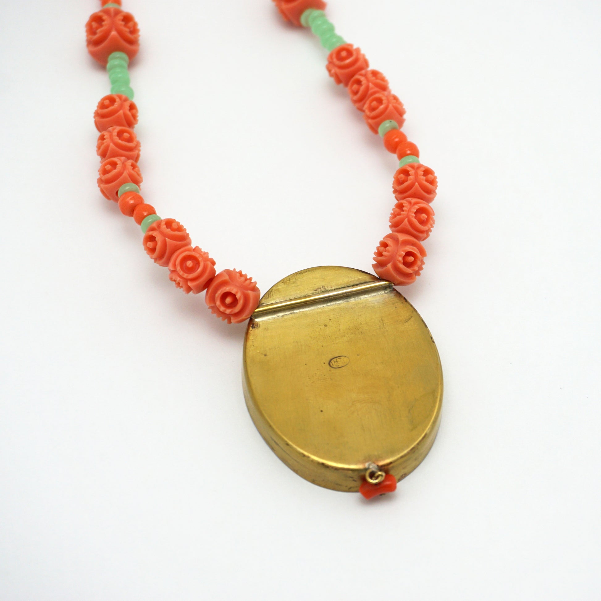 The reverse of a necklace with an oval pendant. The back of the charm is smooth and shiny. The necklace chain is made up of orange and green rose shapes Bakelite beads. 