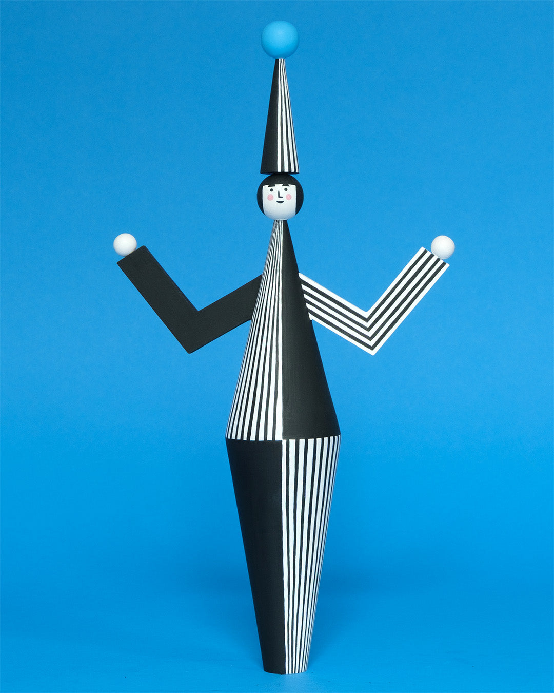 A wooden sculpture with a black and white stripey body with a matching headdress featuring a blue pompom on the top with white and black stripey legs on a plain blue background.