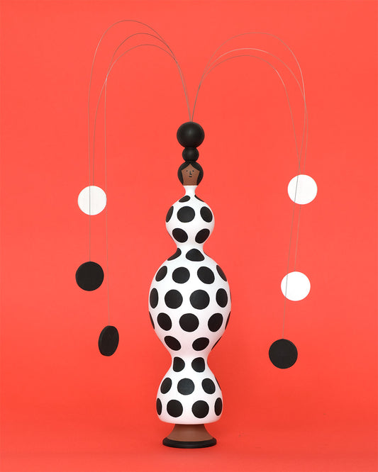 A monochrome sculpture of a sculptural figure with a black and white spotty body with a matching piano wire headdress on a plain orange background.