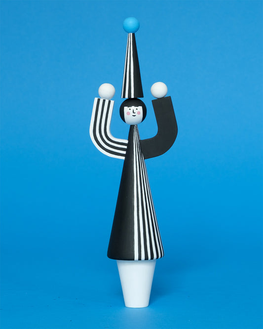 A wooden sculpture with a black and white stripey body with a matching headdress featuring a blue pompom on the top with white legs on a plain blue background.