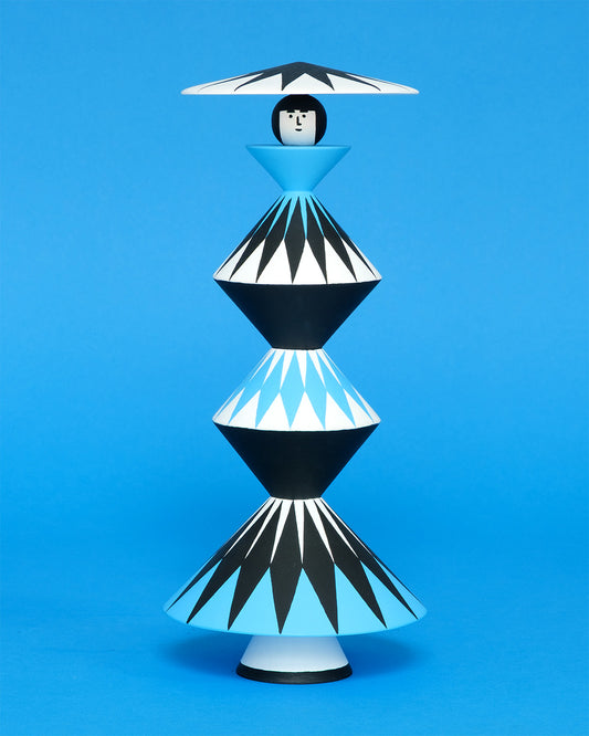 A colourful sculpture with a black, white and blue diamond patterned triangular stacked body with white legs and a matching headdress on a plain blue background.