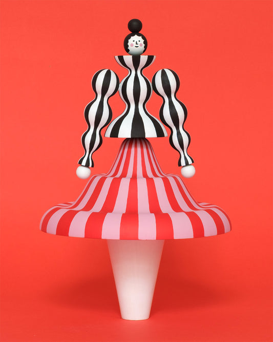 Colourful sculpture of a figure made from a black and white stripey body with a red and pink stripey skirt with white legs on a plain orange background.