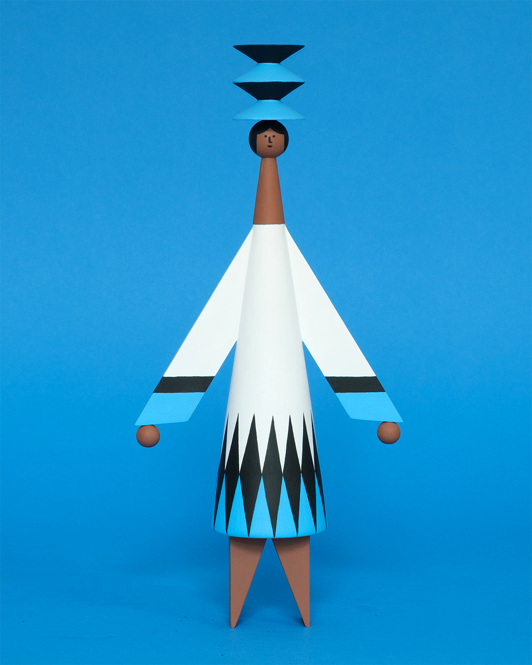 A colourful sculpture with a black, white and blue diamond patterned dress with brown legs and a matching blue and black headdress on a plain blue background.