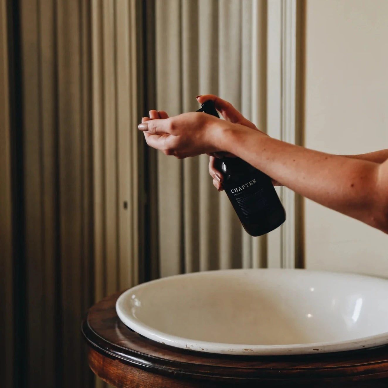 A woman stands by a wooden washstand, applying a product from a dark bottle to her hands. The background features elegant, arched doors and soft curtains, creating a serene, stylish atmosphere.