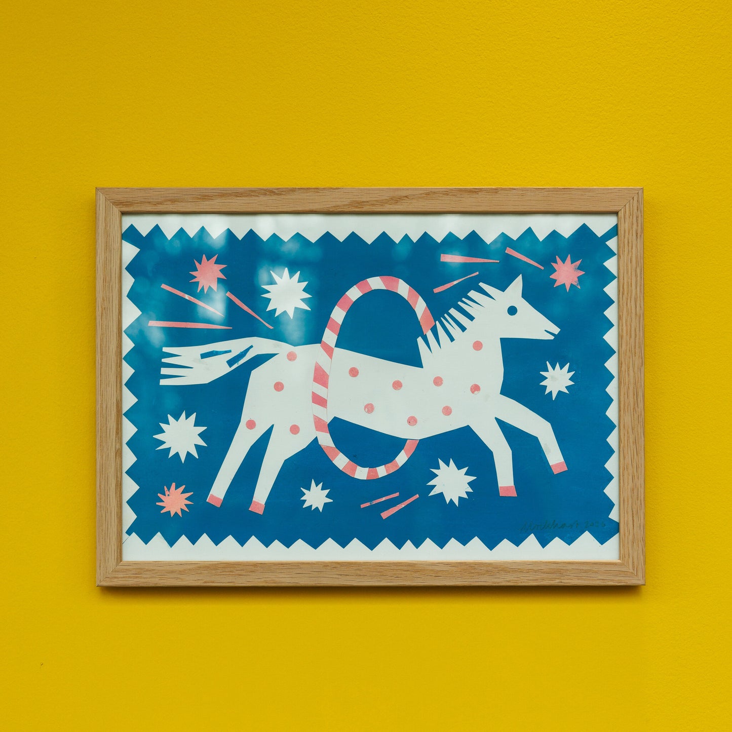 A vibrant artwork featuring a stylised white unicorn with polka dots, jumping through a hoop against a bright blue background. The piece is framed in light wood and set against a bold yellow wall, adding a playful touch to the decor.