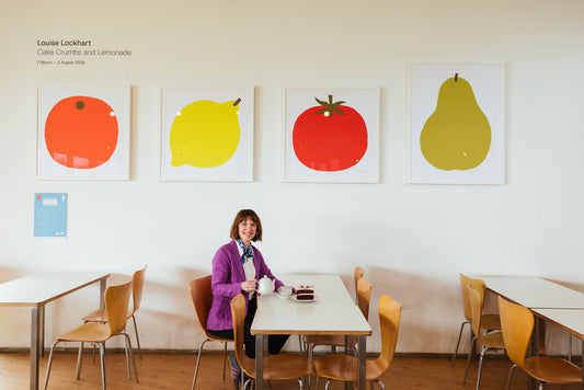 A woman sits at a table in a bright café, wearing a purple jacket and a bow tie. Behind her are five large, colourful fruit prints on the wall. The atmosphere is cheerful and inviting, with wooden furniture and natural light.