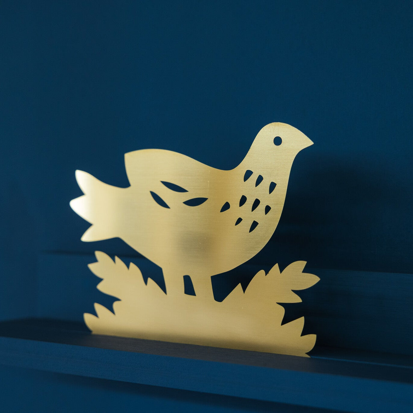 A golden metal bird sculpture perched on a blue shelf, surrounded by stylised leaves. The bird features cut-out details for its eye and wings, creating a delicate and artistic appearance against the contrasting background.