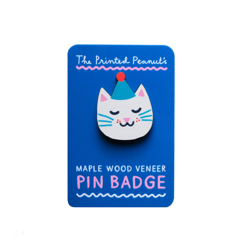 A cute pin badge featuring a stylised cat's face, adorned with a party hat. The badge is made from maple wood veneer and is displayed on a bright blue card that reads "The Printed Peanut's Maple Wood Veneer Pin Badge.