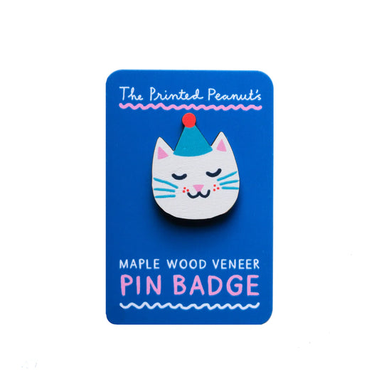 A cute pin badge featuring a stylised cat's face, adorned with a party hat. The badge is made from maple wood veneer and is displayed on a bright blue card that reads "The Printed Peanut's Maple Wood Veneer Pin Badge.