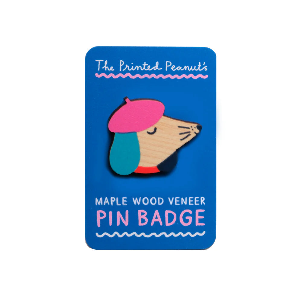 A wooden pin badge featuring a stylised dog wearing a pink beret and blue collar, mounted on a bright blue card. The card reads "The Printed Peanut's Maple Wood Veneer PIN BADGE" in playful white and pink text.