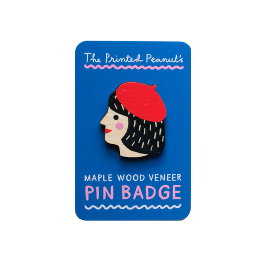 A wooden pin badge featuring a stylised profile of a woman with dark hair and a red beret. The badge is mounted on a bright blue card that reads "The Printed Peanut's Maple Wood Veneer Pin Badge.