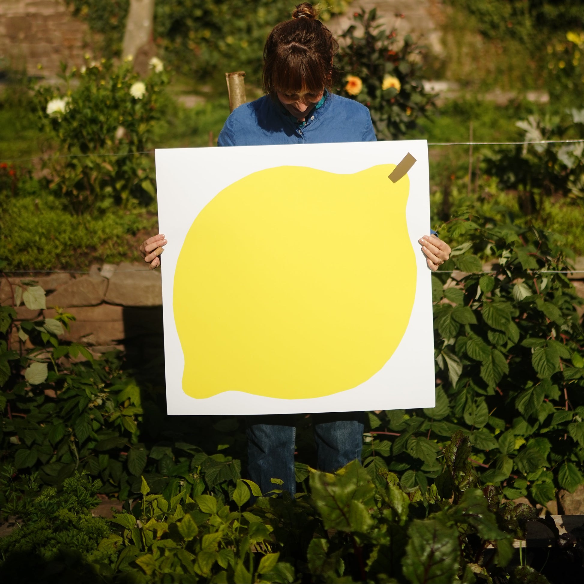 A person holding a simple illustration of a large, bright yellow lemon on a white background. The lemon has a smooth, rounded shape and a small brown stem at the top, emphasising its vibrant colour and fresh appearance.