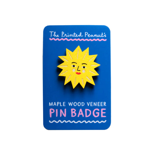 A bright yellow sun-shaped pin badge with a smiling face, mounted on a blue card. The card features the text "The Printed Peanut's" at the top and "MAPLE WOOD VENEER PIN BADGE" in pink at the bottom.