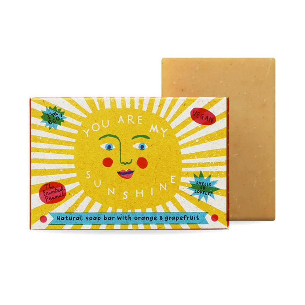 A vibrant soap bar packaging featuring a cheerful sun face with the text "You Are My Sunshine." The box is decorated with bright yellow rays and red accents, highlighting its vegan formula with orange and grapefruit scents. A solid soap bar is visible behind.