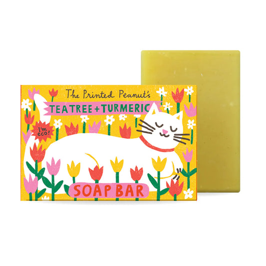 A vibrant soap bar packaging featuring a sleeping white cat surrounded by colourful flowers. The label reads "The Printed Peanut's Tea Tree + Turmeric Soap Bar" with a playful design and eco-friendly message. The soap bar itself is a pale yellow.