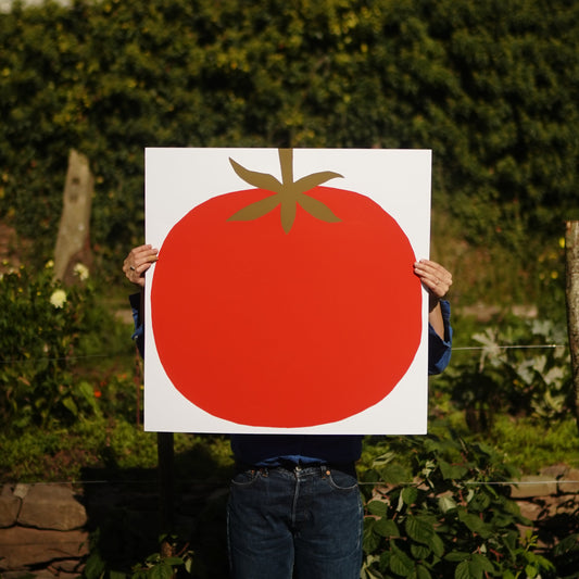 A person holding a simple, stylised illustration of a red tomato on a white background. The tomato is round with a smooth surface and features a small green stem with leaves at the top. The design is minimalistic and bold.