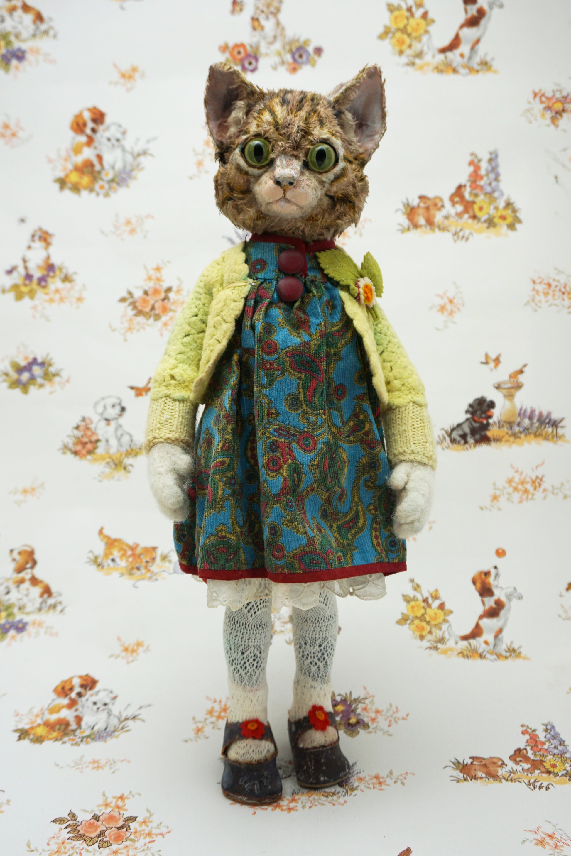 A handmade cat doll with realistic fur, wearing a yellow cardigan, blue patterned dress, and white socks, standing against a white background with a vintage dog and cat pattern.