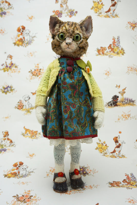 A handmade cat doll with realistic fur, wearing a yellow cardigan, blue patterned dress, and white socks, standing against a white background with a vintage dog and cat pattern.