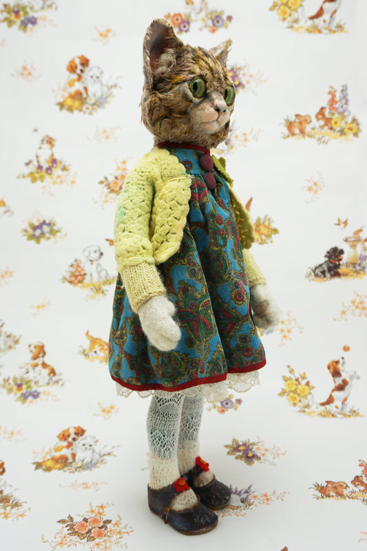A handmade cat doll with green eyes, dressed in a yellow cardigan, blue patterned dress, white socks, and dark shoes, standing against a white background with a vintage-style dog and cat pattern.