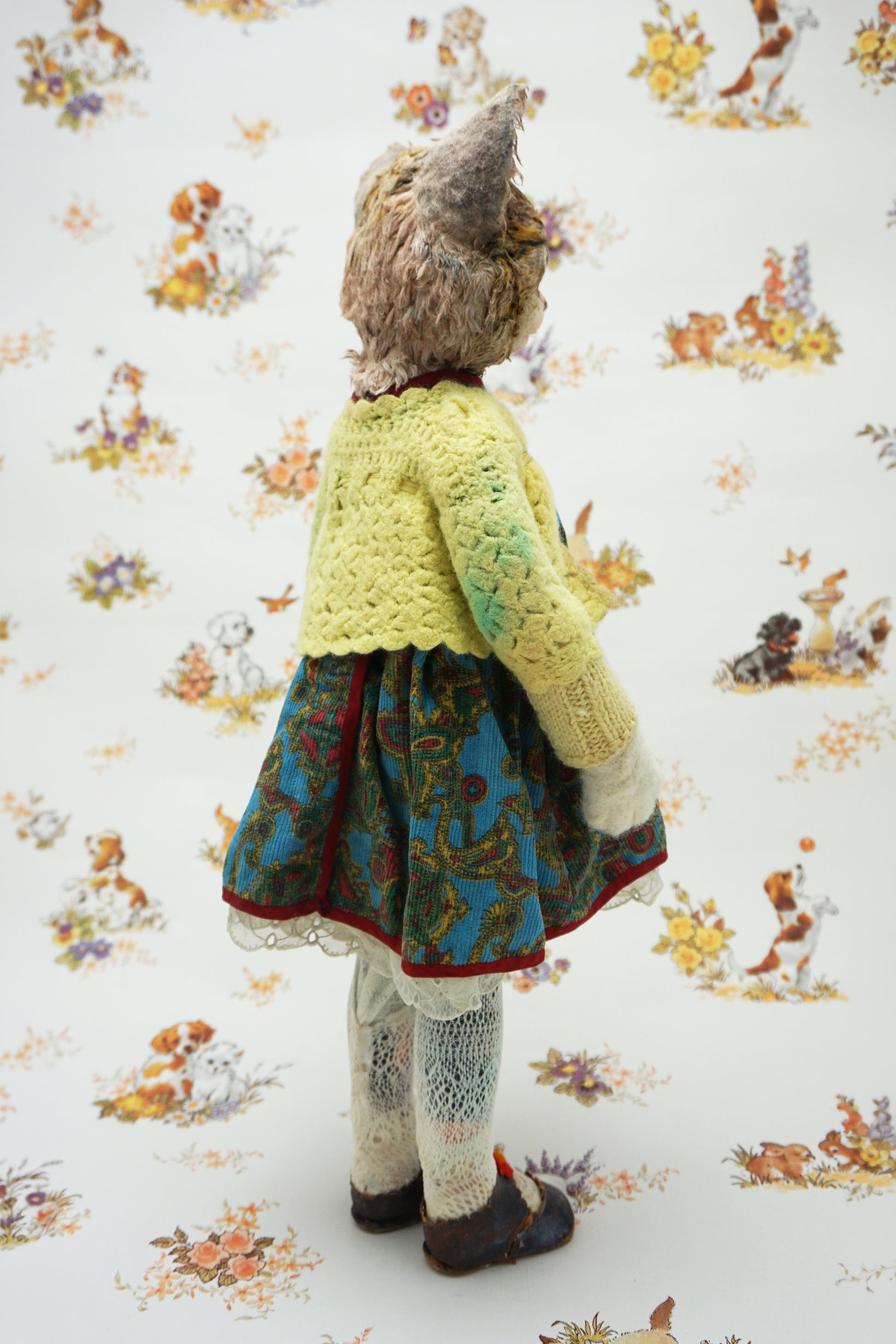 A vintage-style doll or collectible figure shown from the back, wearing a yellow crocheted cardigan, a blue patterned skirt, and white lace socks, standing against a white floral wallpaper background.