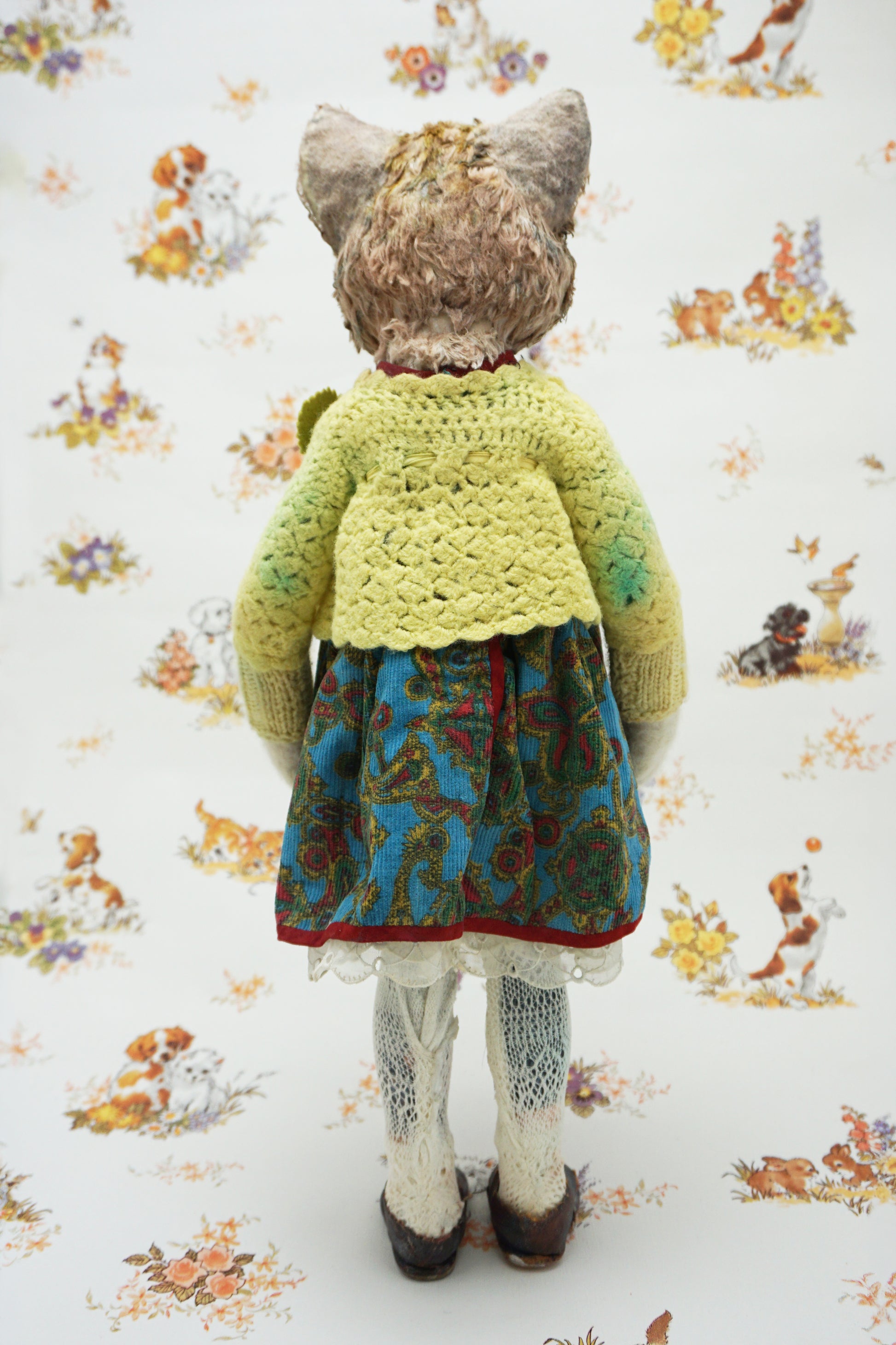 A vintage-style doll viewed from behind, wearing a yellow-green crochet cardigan, a blue and green floral skirt, white lace-trimmed tights, and dark shoes, standing against a white floral wallpaper background.