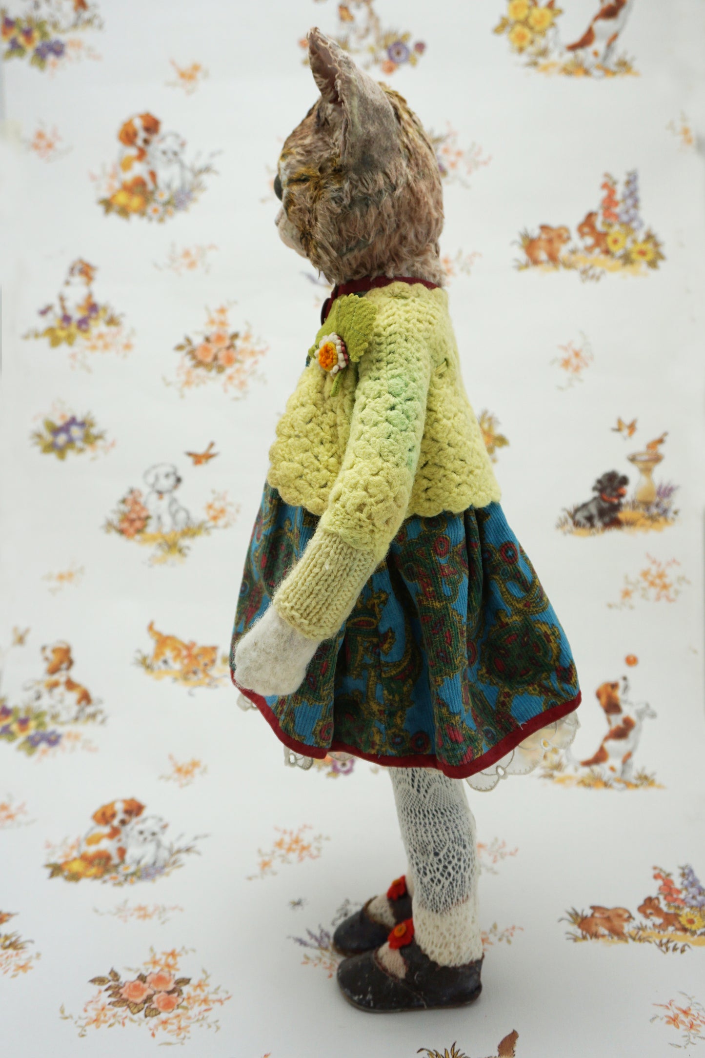 A handmade plush cat doll shown from the back, wearing a light green crocheted cardigan, a blue and green patterned dress, white socks, and dark shoes, set against a white background with a vintage-style animal print.